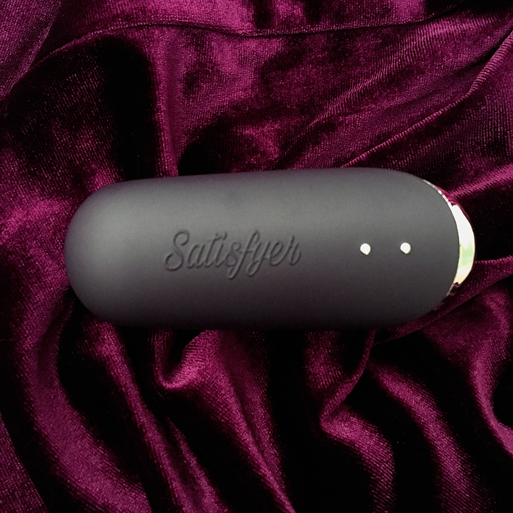 above photo of dark purple Satisfyer Pro Traveler on red velvet background. The name "satisfyer" is embelished on the side, two metal magnet charging buttons on the bottom