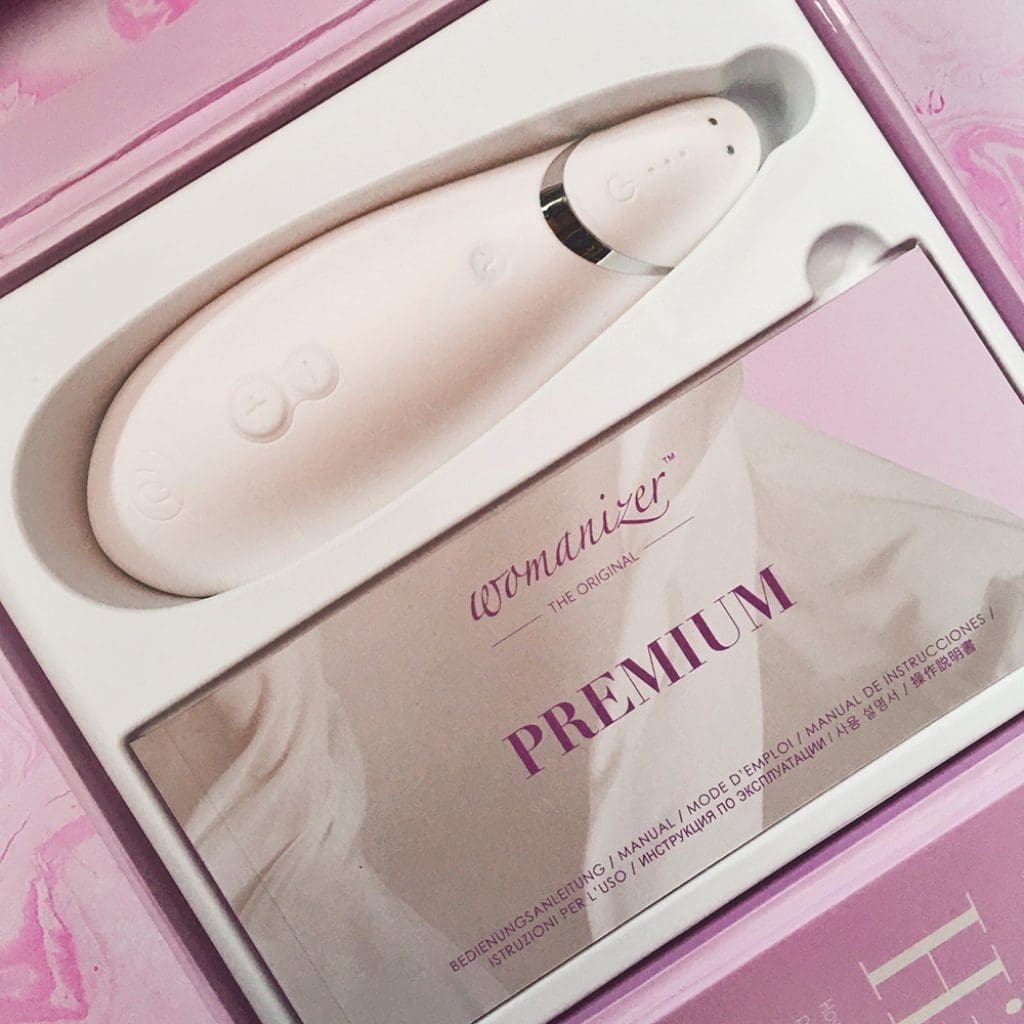 photograph showing inside of Womanizer Premium packaging with white silicone oval shaped air-pulse toy and instruction manual on pink background