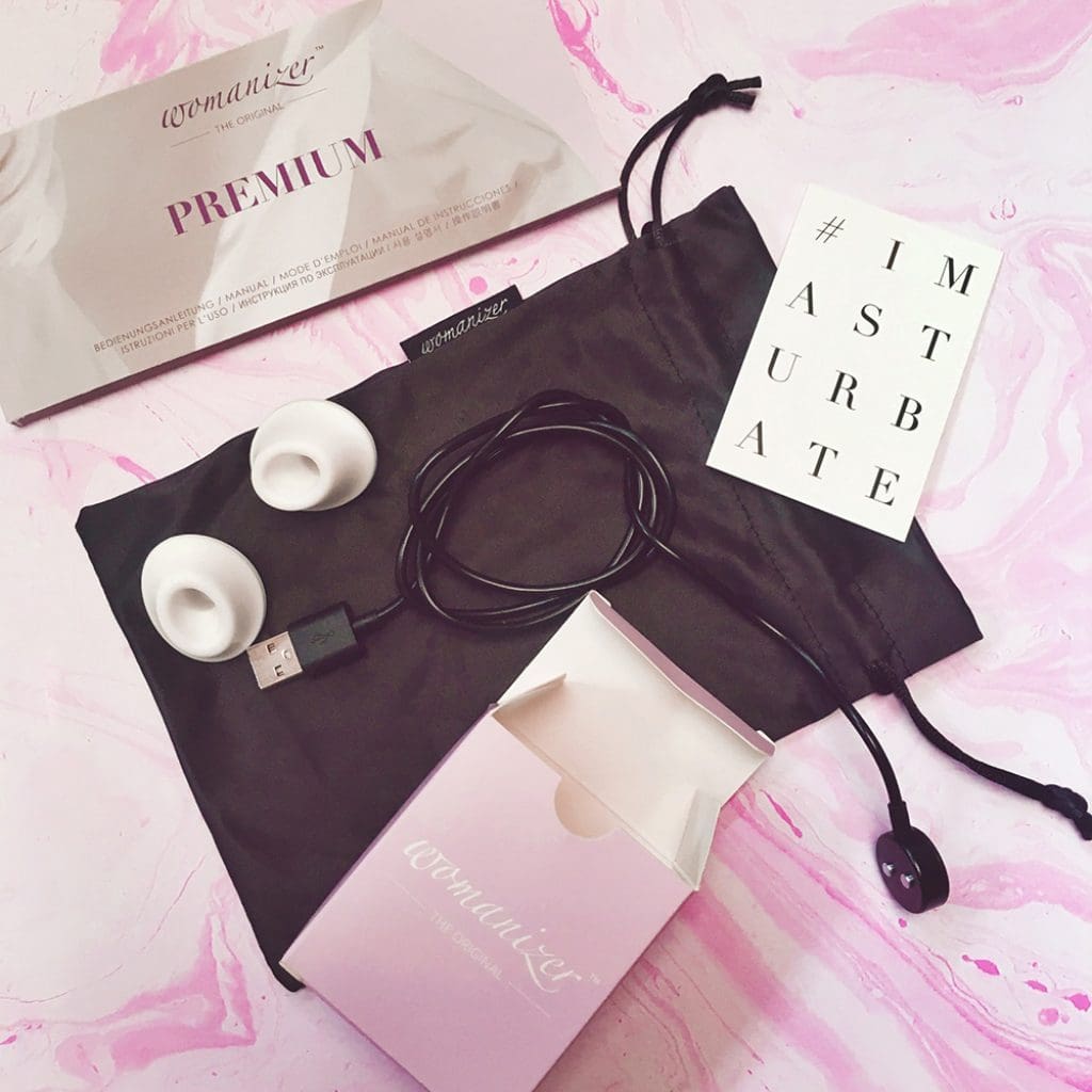 photograph of contents of Womanizer Premium packaging spread out - paper manual, black cloth drawstring bag, charging cable, two nozzles, and white card that reads # I Masturbate