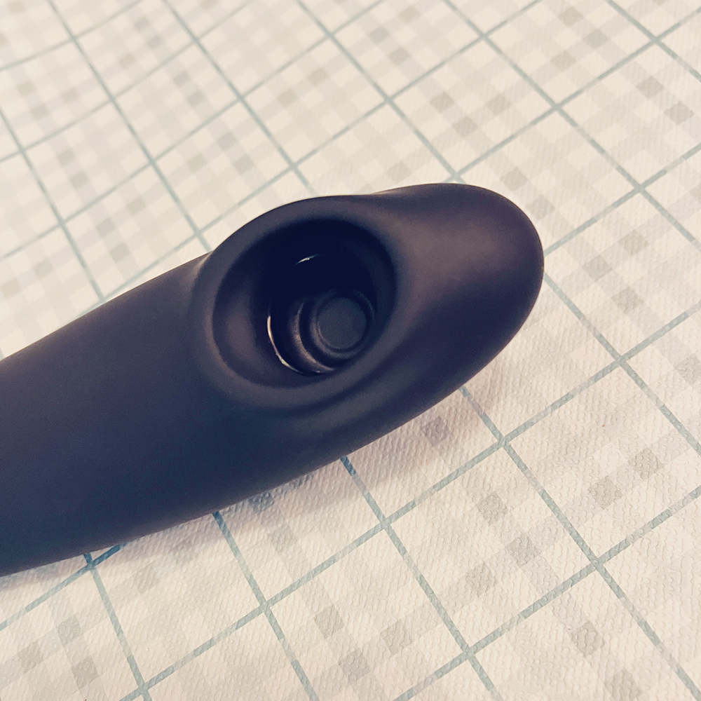 Photograph of slate grey insertable air-pulse toy showing the large shallow oval shaped nozzle, several ridges where toy components join together, and silicone membrane covering the bottom of the well