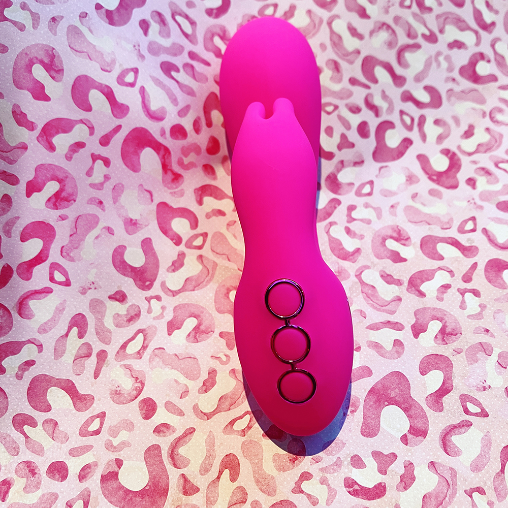 photo of hot pink U-shaped dual stimulator on its back showing 3 circular buttons at bottom of U bend, top stimulator arm has two "bunny ear" prongs, bottom stimulator arm is oval shaped