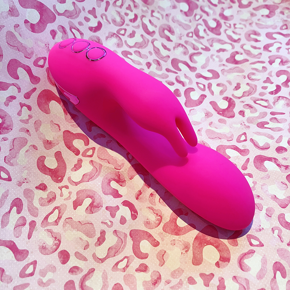 photo of hot pink dual stimulator vibe on its back turned to the side to get a better look at the prong "bunny ears" clitoral arm and oval internal arm