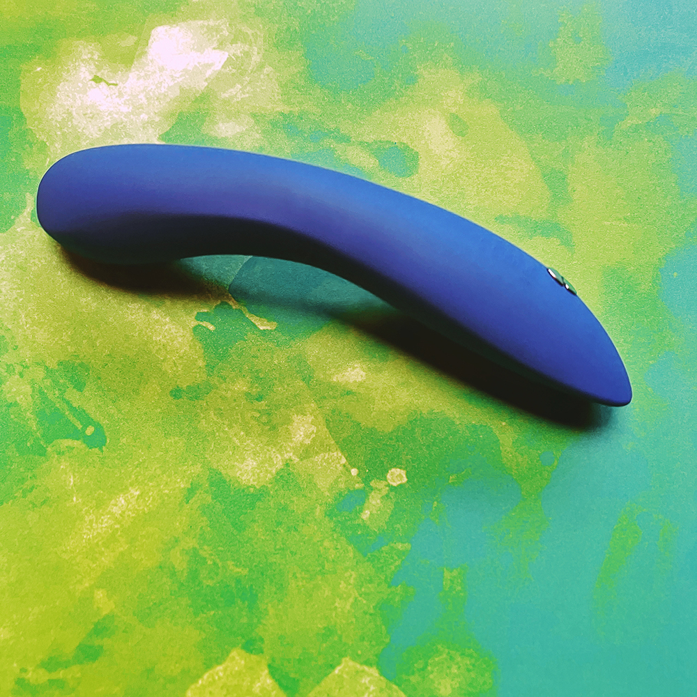 photograph of light blue abstract cylindrical vibrator on green background. Vibrator is on it's face showing a slight doming bend. metal magnetic charging pins on the back of the handle