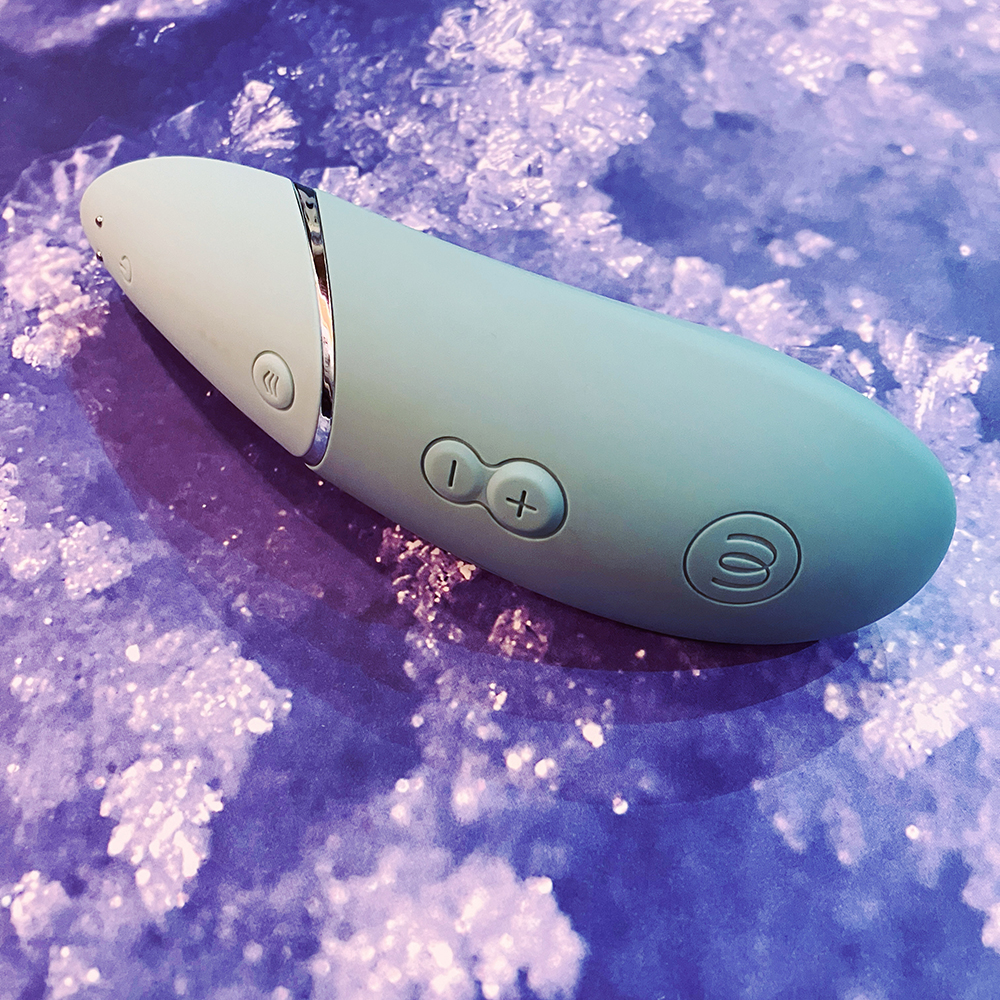 Photograph of mint green long oval shaped clitoral Sex toy with silver accents. Photograph shows two-pin magnetic charging port at bottom of handle, power button, depth control button, minus and plus intensity buttons, and Womanizer logo